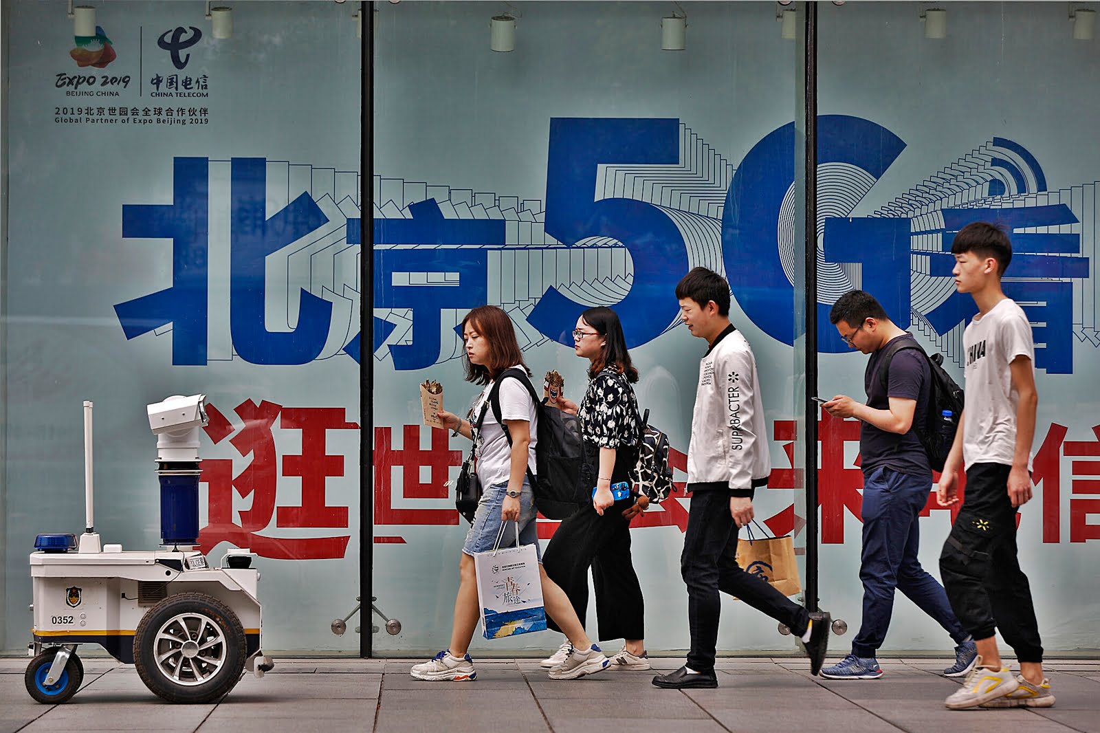 Photo Credit: Sebuah robot polisi yang dipasang dengan kamera pengintai menyaksikan orang-orang berjalan melewati iklan jaringan 5G di sebuah distrik perbelanjaan di Beijing, Rabu, 15 Mei 2019. AP/Andy Wong